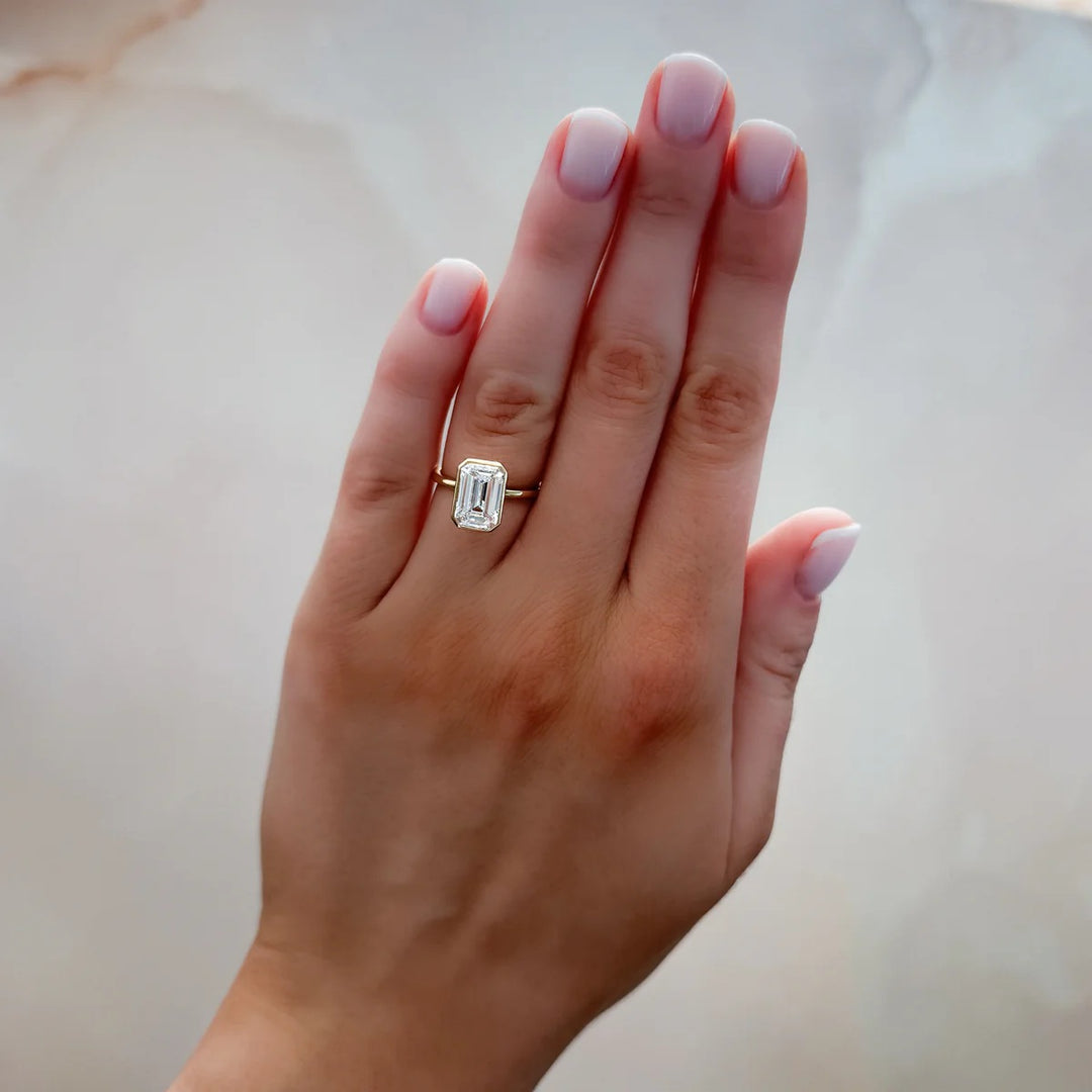 Woman wearing 2.50ct emerald cut bezel set diamond ring on finger, 14k yellow gold thin band, showing modern style