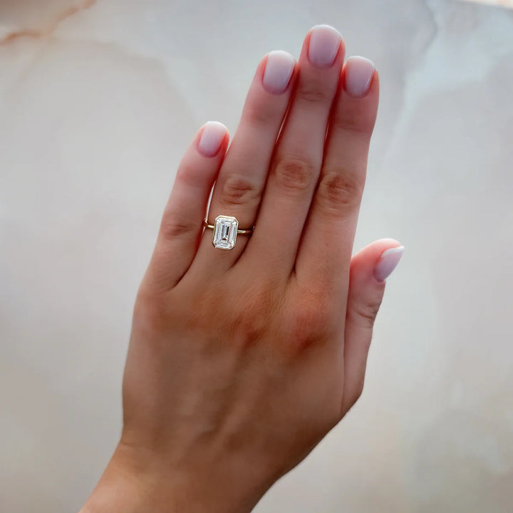 Woman wearing 2.50ct emerald cut bezel set diamond ring on finger, 14k yellow gold thin band, showing modern style
