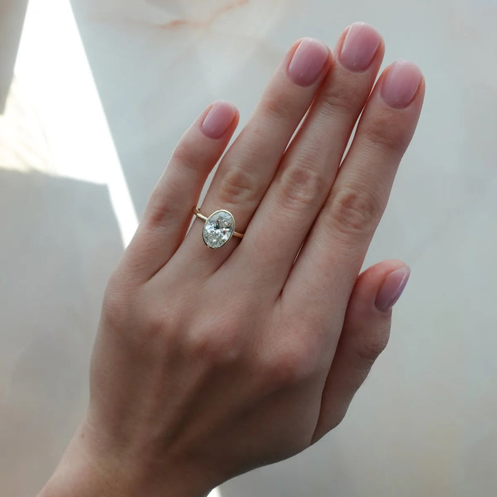 Woman wearing 2.50ct oval bezel set diamond ring on finger, 14k yellow gold thin band, showing modern style